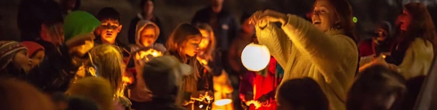 Photo of students gathered around a teacher who is holding a lantern and telling a story in the evening during Lantern Walk Header Festival