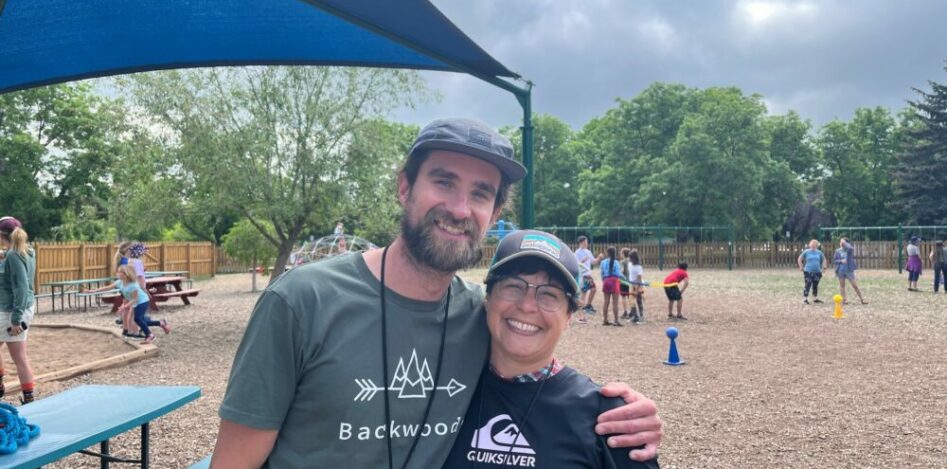 Photo of two teachers on the playground, standing side by side with their arms around each other, smiling at the camera