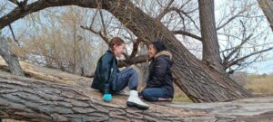 Photo of two students sitting on top of a fallen cottonwood tree in the woods, facing each other and smiling